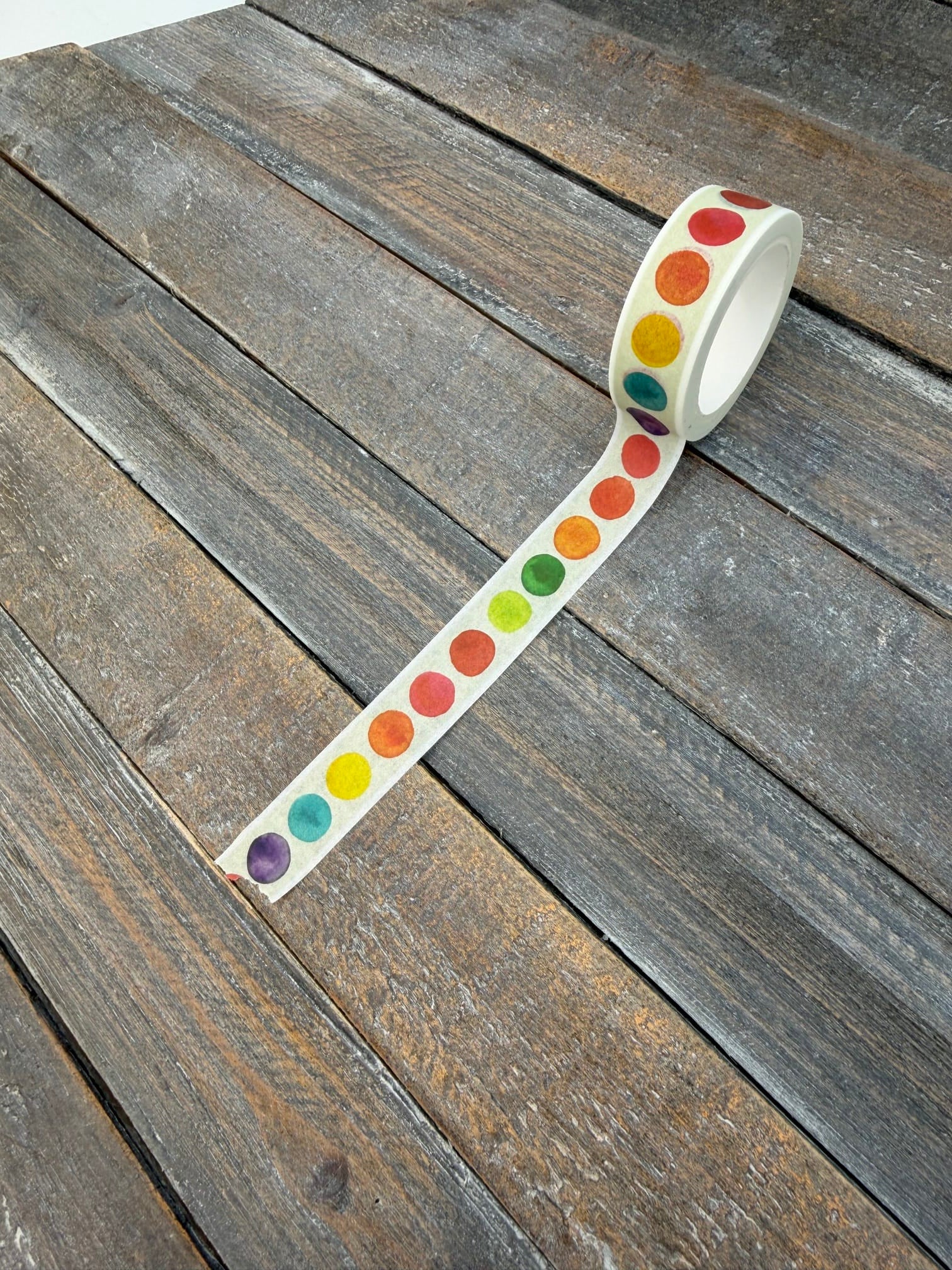 A roll of Color Theory washi tape partly unspooled on weathered grey wood, showing the full spectrum of watercolor dots running along the cream tape.