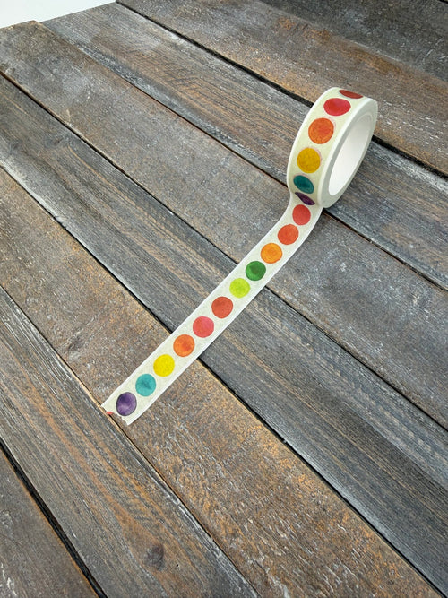 A roll of Color Theory washi tape partly unspooled on weathered grey wood, showing the full spectrum of watercolor dots running along the cream tape.