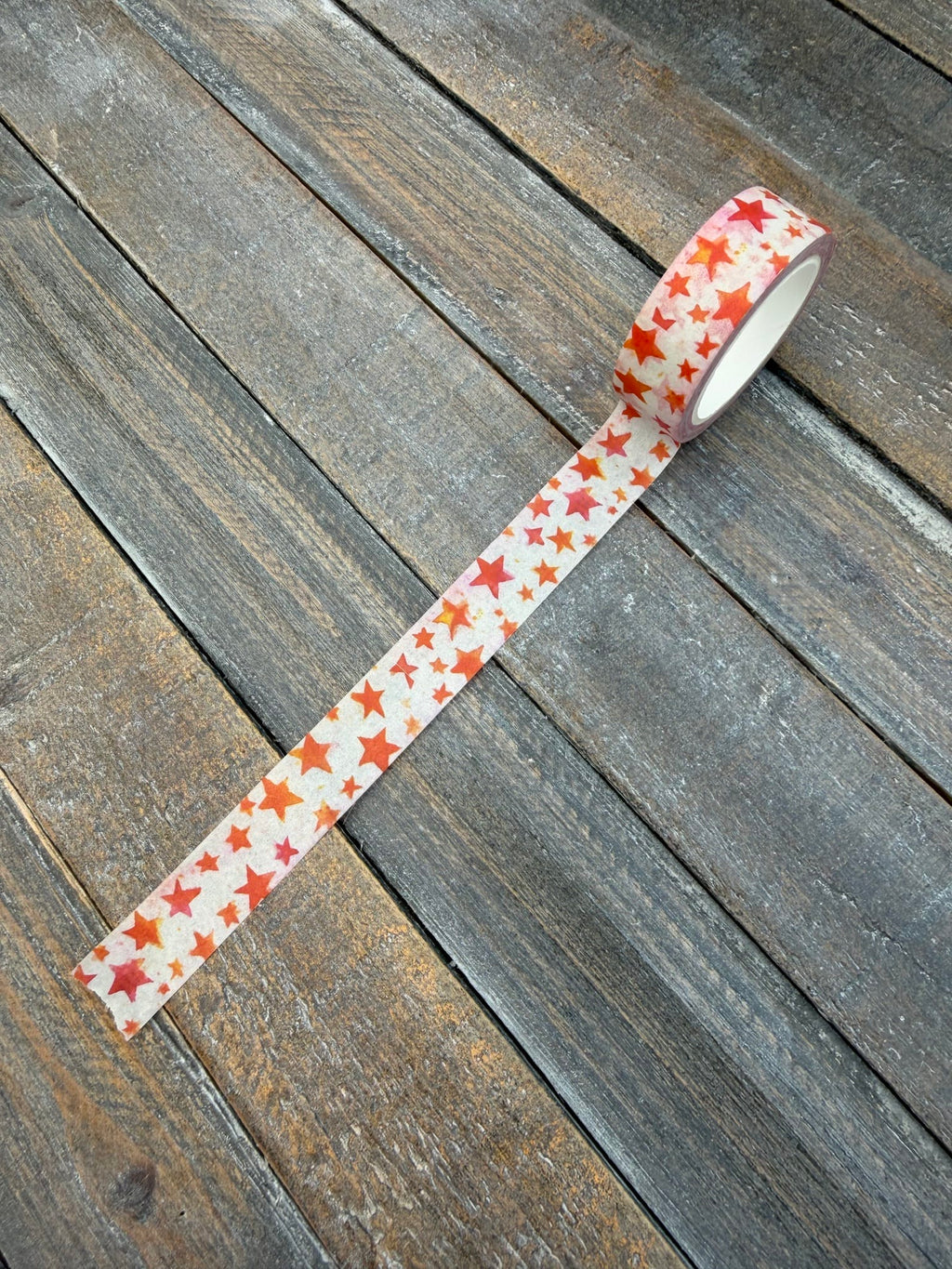 A roll of Seeing Stars washi tape partly unspooled on weathered grey wood, the white tape scattered with hand-painted watercolor stars in red, orange, coral and pink.