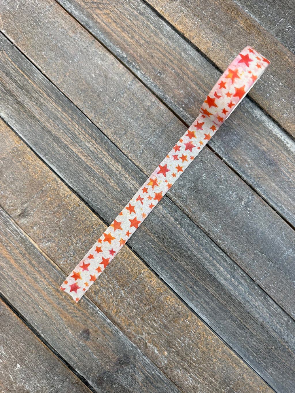 A strip of white washi tape scattered with hand-painted watercolor stars in red, orange, coral and pink, laid diagonally across weathered grey barn wood.