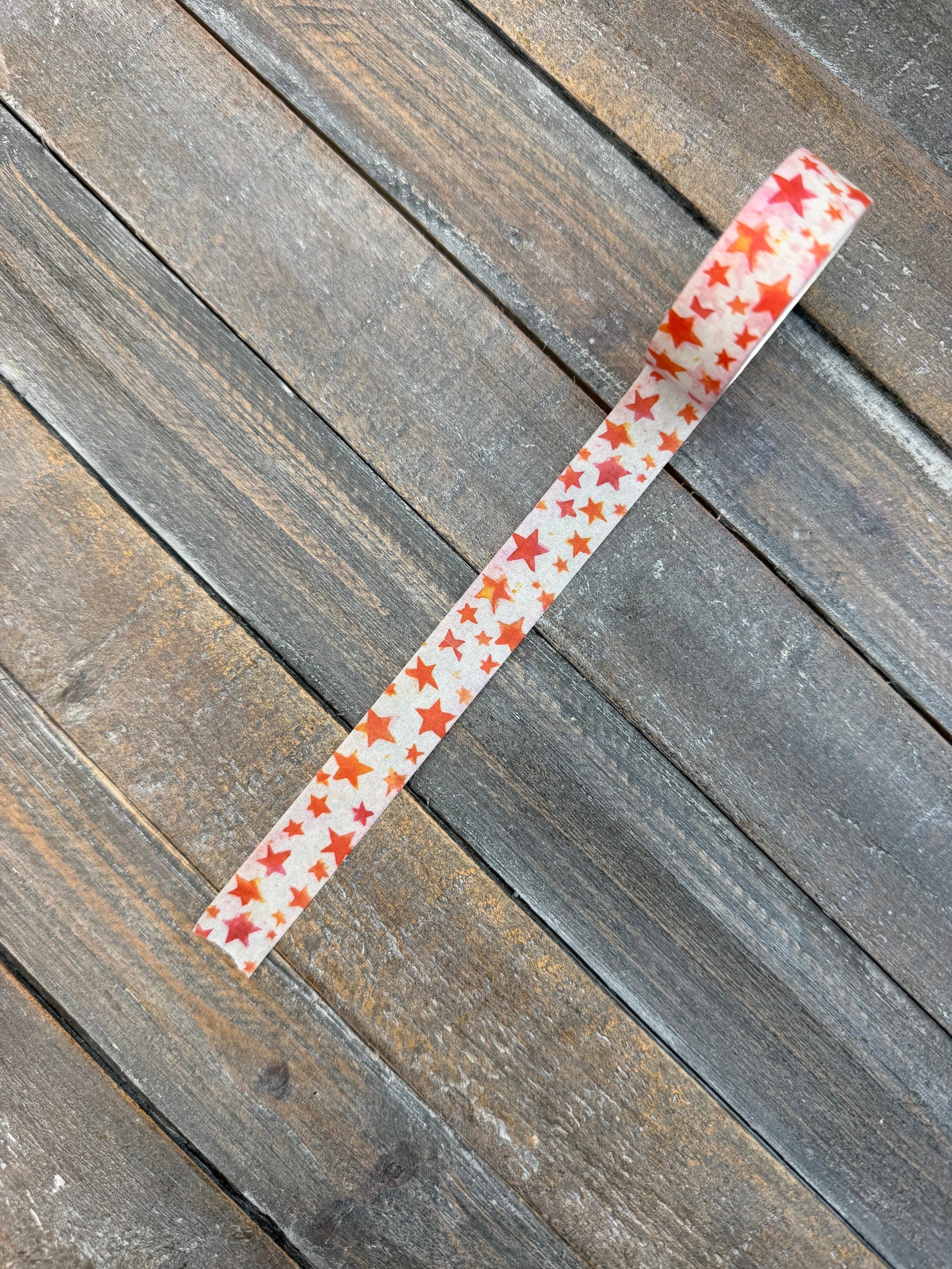 A strip of white washi tape scattered with hand-painted watercolor stars in red, orange, coral and pink, laid diagonally across weathered grey barn wood.