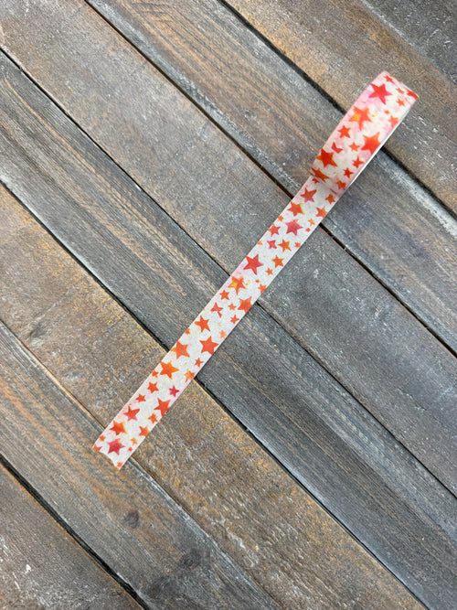 A strip of white washi tape scattered with hand-painted watercolor stars in red, orange, coral and pink, laid diagonally across weathered grey barn wood.