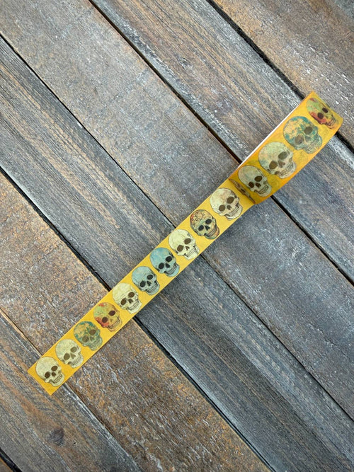 The Bone Parade Washi Tape — unrolled roll of golden-orange washi tape with a repeating weathered skull motif, laid diagonally across a weathered wood surface